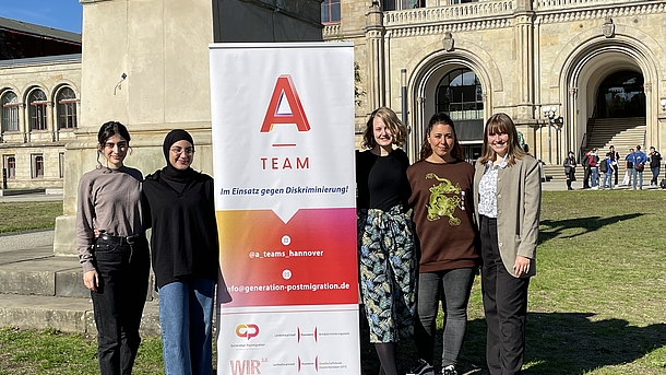 Vier Studentinnen stehen vor dem Hauptgebäude der Universität Hannover mit Einem A-Team-Aufsteller